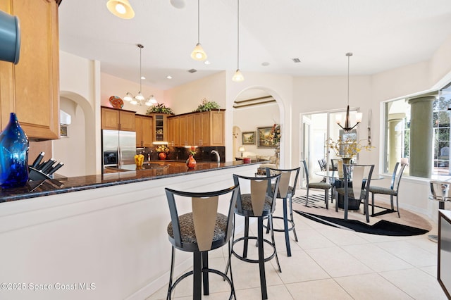 kitchen with a chandelier, arched walkways, stainless steel refrigerator with ice dispenser, brown cabinetry, and glass insert cabinets