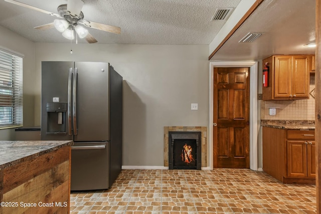 kitchen with ceiling fan, stainless steel refrigerator with ice dispenser, a textured ceiling, decorative backsplash, and dark stone counters