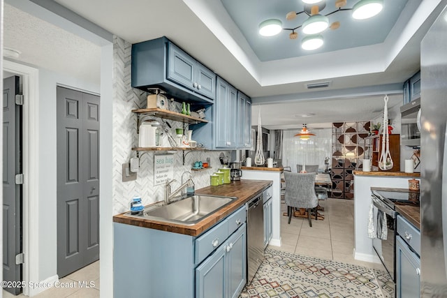 kitchen featuring a tray ceiling, light tile patterned floors, stainless steel appliances, butcher block counters, and a sink