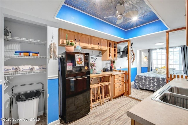 kitchen featuring black refrigerator, sink, ceiling fan, a raised ceiling, and light wood-type flooring