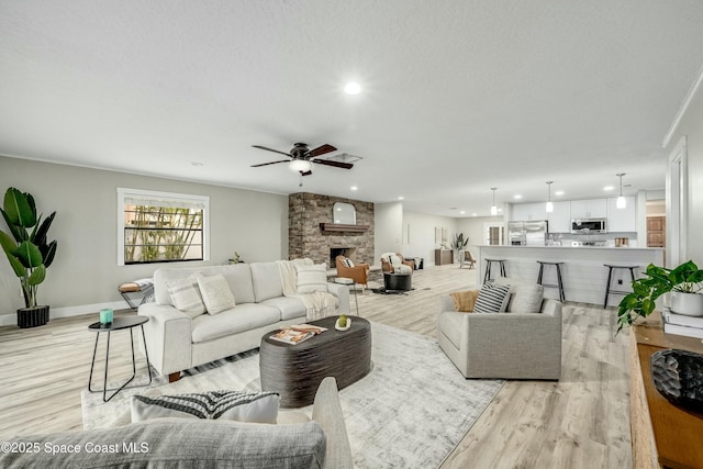 living room with baseboards, a stone fireplace, light wood-style flooring, and a ceiling fan