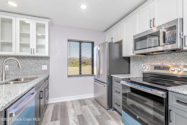 kitchen with a sink, white cabinets, appliances with stainless steel finishes, light stone countertops, and glass insert cabinets