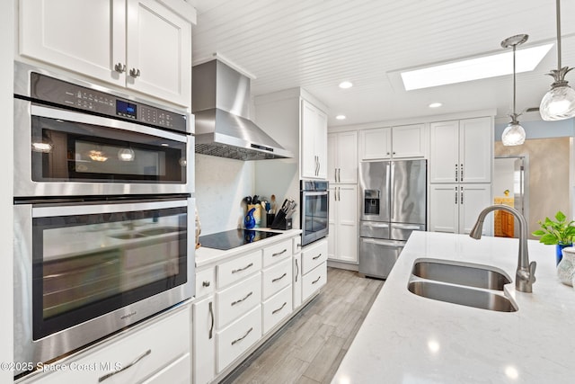 kitchen with appliances with stainless steel finishes, wall chimney range hood, white cabinetry, a skylight, and a sink