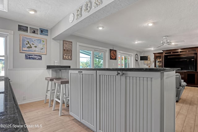 kitchen with light wood-style flooring, a ceiling fan, a textured ceiling, a breakfast bar area, and wainscoting