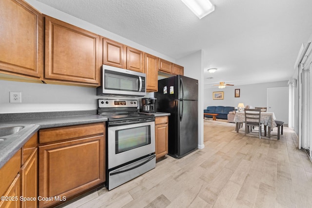 kitchen with a textured ceiling, light wood-style flooring, appliances with stainless steel finishes, brown cabinetry, and dark countertops