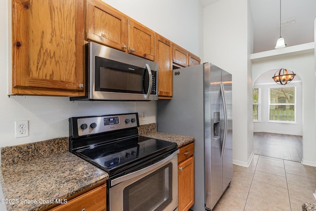 kitchen featuring light tile patterned floors, brown cabinetry, appliances with stainless steel finishes, dark stone countertops, and pendant lighting