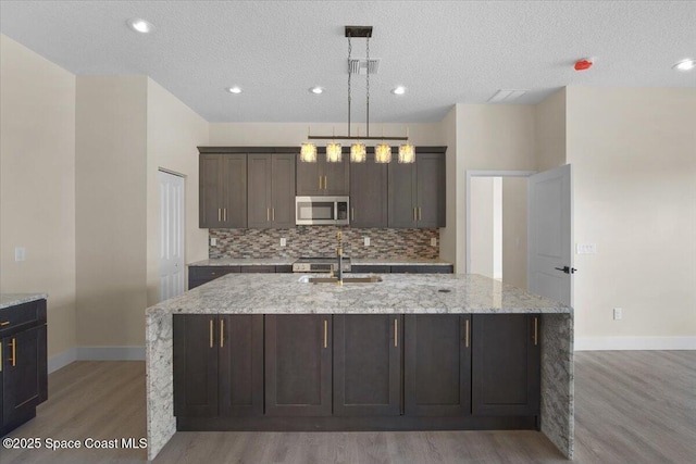 kitchen featuring a kitchen island with sink, a sink, decorative backsplash, stainless steel microwave, and pendant lighting