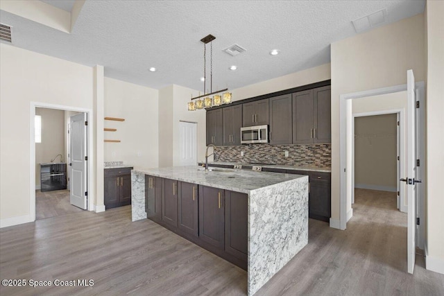 kitchen with decorative light fixtures, stainless steel microwave, a sink, dark brown cabinets, and an island with sink