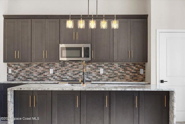 kitchen featuring light stone counters, tasteful backsplash, stainless steel microwave, hanging light fixtures, and dark brown cabinetry