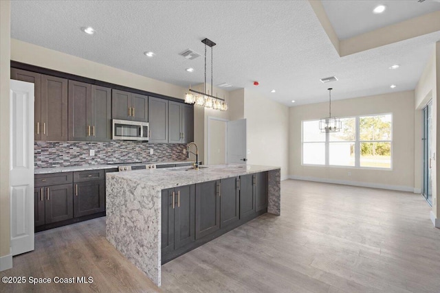 kitchen featuring a kitchen island with sink, pendant lighting, stainless steel microwave, and dark brown cabinets