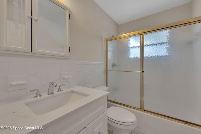 bathroom with bath / shower combo with glass door, toilet, a wainscoted wall, vanity, and tile walls