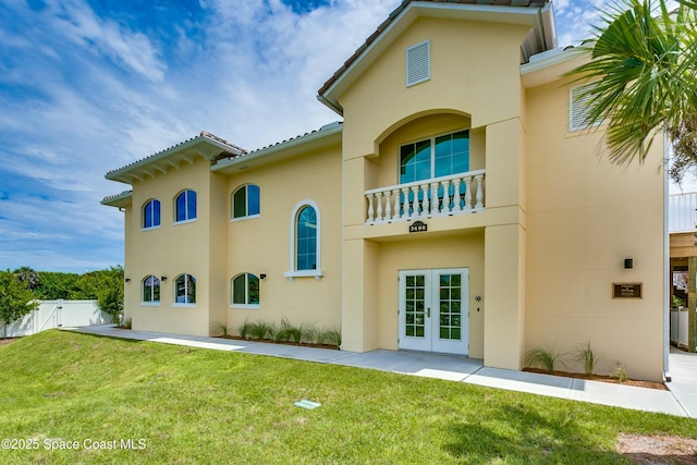 rear view of house featuring french doors, stucco siding, a lawn, fence, and a balcony