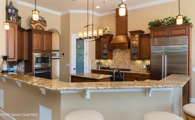 kitchen with hanging light fixtures, appliances with stainless steel finishes, brown cabinetry, and glass insert cabinets