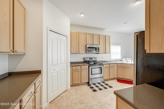 kitchen with light tile patterned floors, light brown cabinets, stainless steel appliances, a sink, and dark countertops