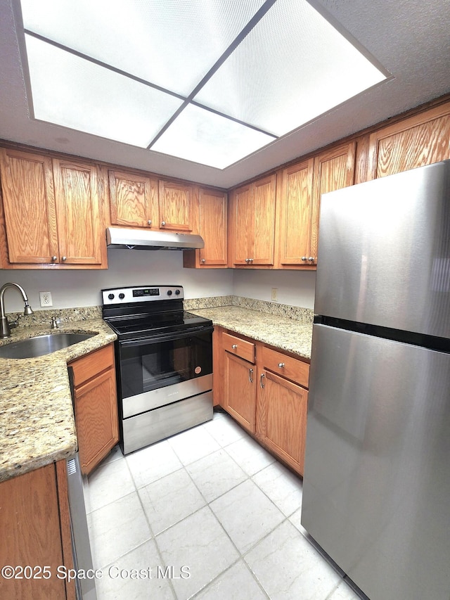 kitchen with brown cabinetry, appliances with stainless steel finishes, light stone countertops, under cabinet range hood, and a sink