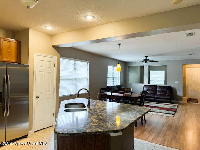 kitchen featuring visible vents, a center island with sink, a sink, dark stone countertops, and stainless steel fridge