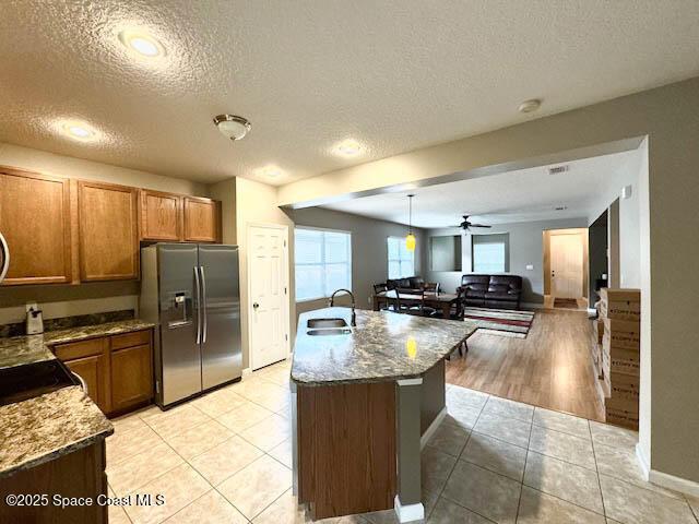 kitchen with stainless steel fridge with ice dispenser, an island with sink, light tile patterned floors, brown cabinets, and a sink