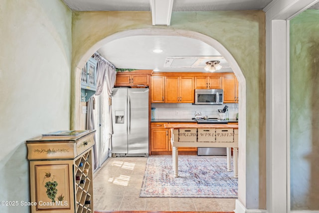 2109 Indian River Dr, Cocoa, FL 32922 - kitchen featuring stainless steel appliances, arched walkways, tasteful backsplash, brown cabinetry, and light tile patterned floors
