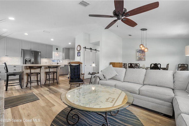 living room featuring a barn door, visible vents, a ceiling fan, vaulted ceiling, and light wood-type flooring