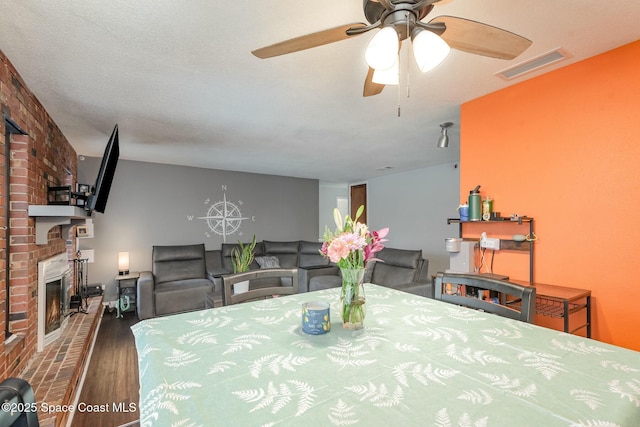 dining area featuring a brick fireplace, ceiling fan, visible vents, and wood finished floors