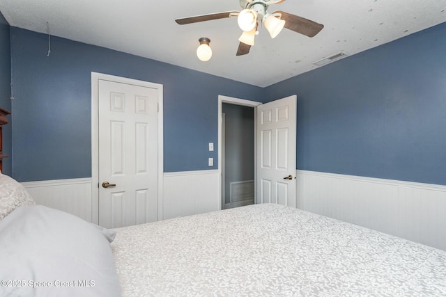 bedroom with a wainscoted wall, attic access, visible vents, and a ceiling fan