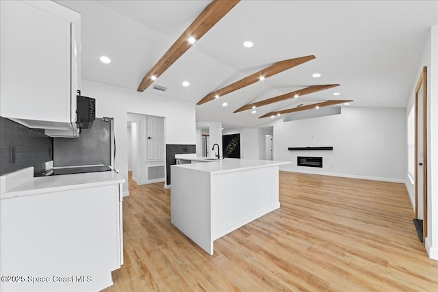 kitchen with a glass covered fireplace, lofted ceiling with beams, light wood-style floors, white cabinetry, and a sink