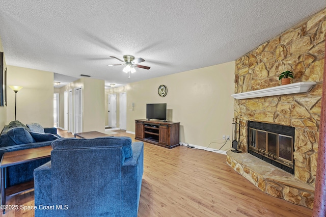 living area featuring light wood finished floors, ceiling fan, a stone fireplace, and a textured ceiling