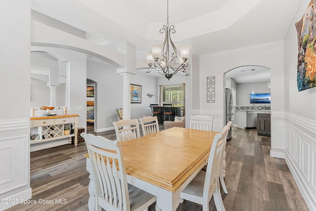 dining space featuring arched walkways, dark wood-style flooring, wainscoting, a tray ceiling, and ornate columns