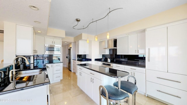 kitchen with dark countertops, appliances with stainless steel finishes, white cabinetry, a sink, and wall chimney range hood