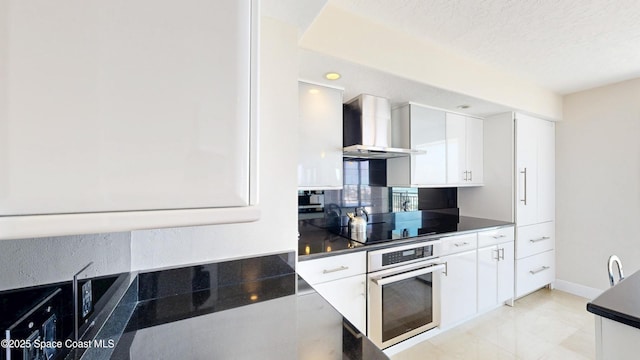 kitchen featuring dark countertops, wall chimney range hood, stainless steel oven, and white cabinets