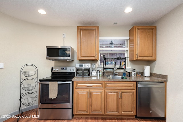 kitchen featuring wood finished floors, a sink, appliances with stainless steel finishes, brown cabinetry, and dark countertops