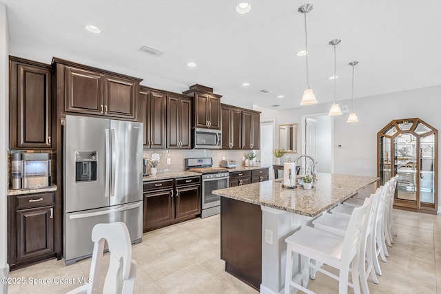 kitchen with light stone counters, dark brown cabinets, appliances with stainless steel finishes, decorative backsplash, and a center island with sink