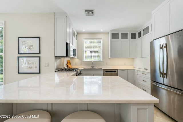 kitchen featuring a peninsula, a sink, visible vents, appliances with stainless steel finishes, and glass insert cabinets