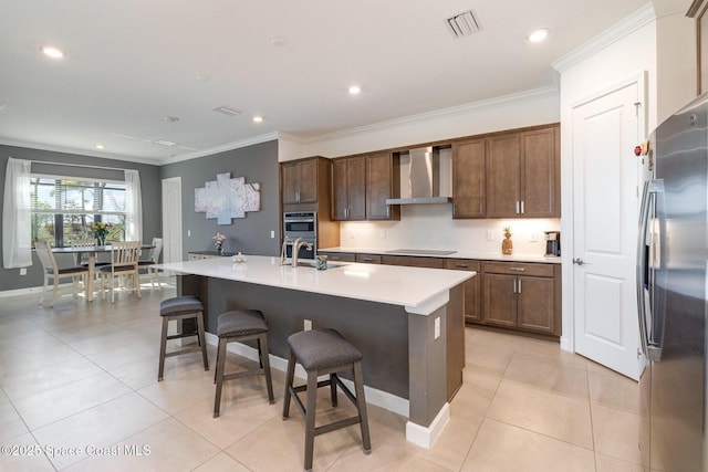 kitchen featuring a breakfast bar, an island with sink, appliances with stainless steel finishes, crown molding, and wall chimney range hood