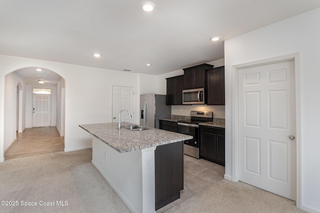kitchen featuring appliances with stainless steel finishes, a sink, an island with sink, arched walkways, and light carpet