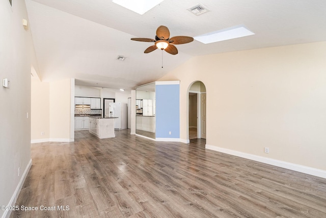 unfurnished living room with ceiling fan, light wood-style flooring, visible vents, baseboards, and lofted ceiling with skylight