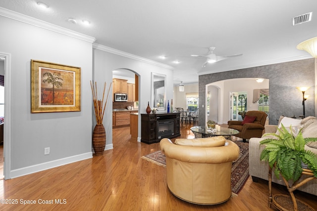 living area featuring baseboards, visible vents, arched walkways, light wood-style flooring, and crown molding