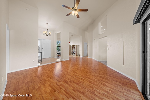 unfurnished living room with light wood-type flooring, a towering ceiling, baseboards, and ceiling fan with notable chandelier