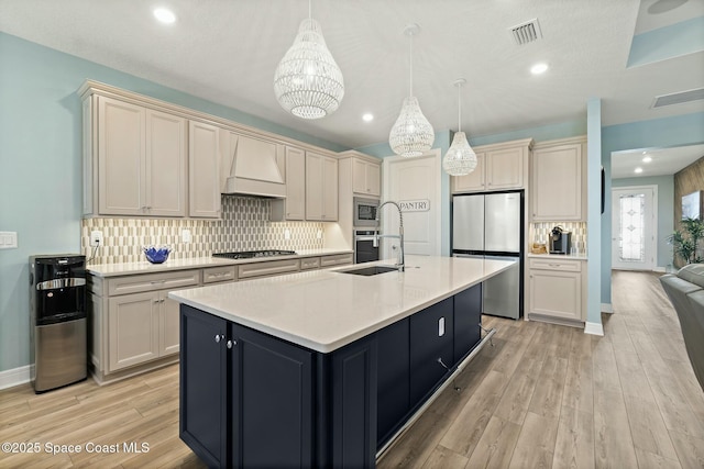 kitchen with stainless steel appliances, custom range hood, visible vents, light wood-style floors, and an island with sink