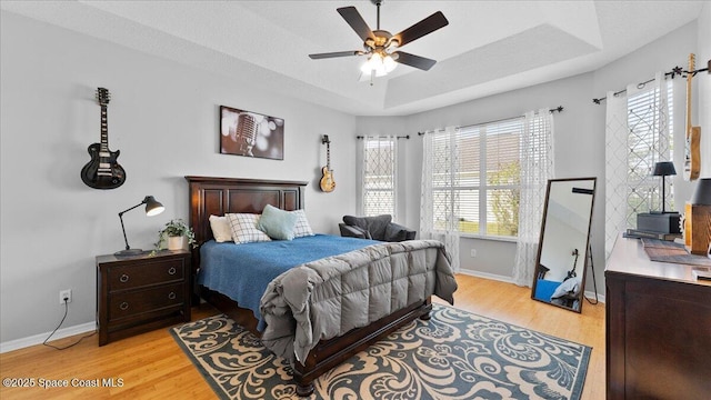 bedroom with light wood finished floors, baseboards, multiple windows, and a tray ceiling