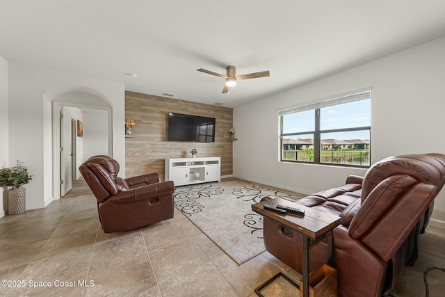 living room with light tile patterned floors, an accent wall, wood walls, a ceiling fan, and baseboards