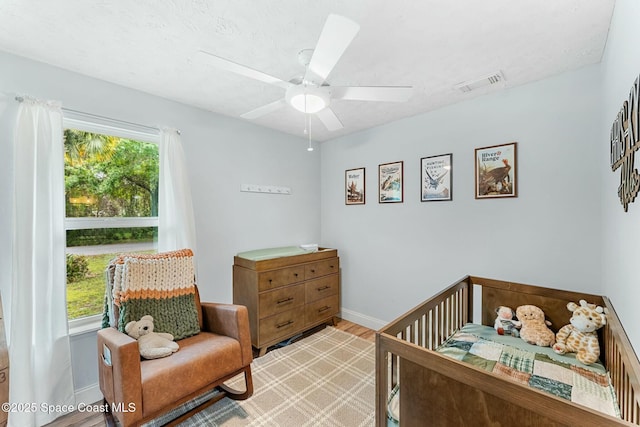 bedroom featuring visible vents, light wood-style flooring, a ceiling fan, a nursery area, and baseboards