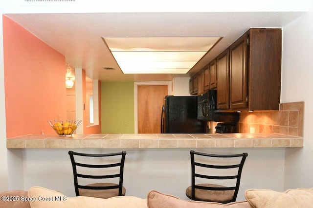 kitchen with a peninsula, a kitchen bar, tile counters, and black fridge