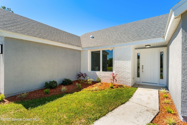 property entrance featuring a yard, brick siding, roof with shingles, and stucco siding