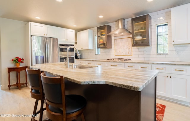 kitchen with stainless steel appliances, a sink, light wood-type flooring, backsplash, and wall chimney exhaust hood