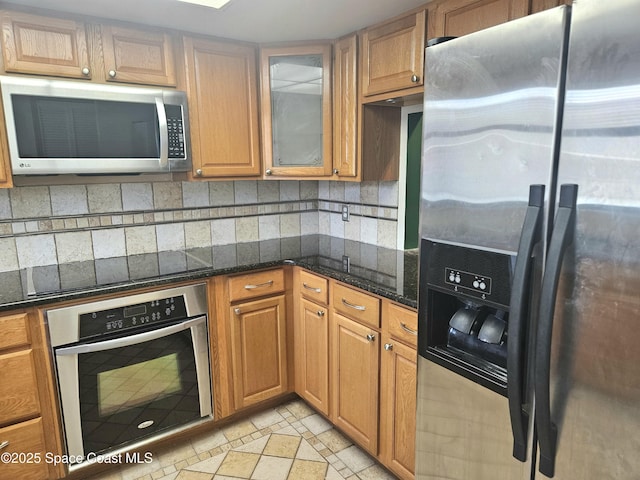 kitchen featuring brown cabinets, stainless steel appliances, decorative backsplash, glass insert cabinets, and dark stone counters