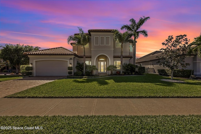 mediterranean / spanish home with a garage, a tiled roof, decorative driveway, a lawn, and stucco siding