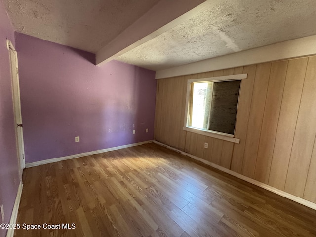 empty room featuring a textured ceiling, baseboards, wood finished floors, and beamed ceiling