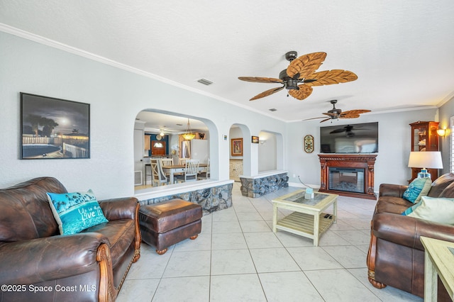 tiled living area featuring arched walkways, visible vents, a ceiling fan, ornamental molding, and a glass covered fireplace