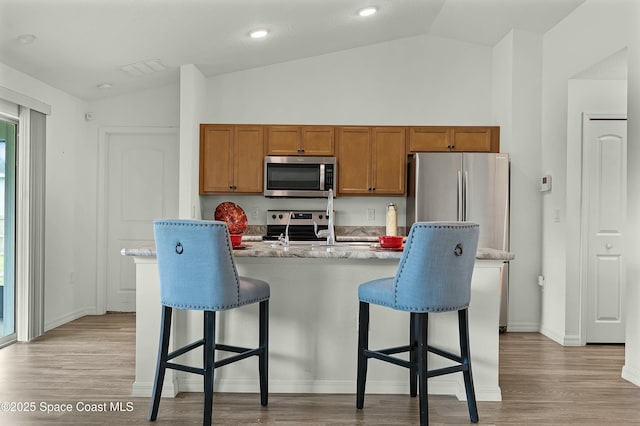 kitchen featuring light wood finished floors, stainless steel appliances, brown cabinetry, vaulted ceiling, and a kitchen breakfast bar